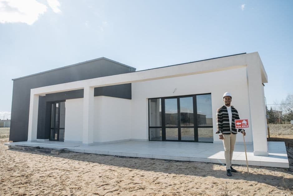 A realtor stands with a for sale sign in front of a modern house.
